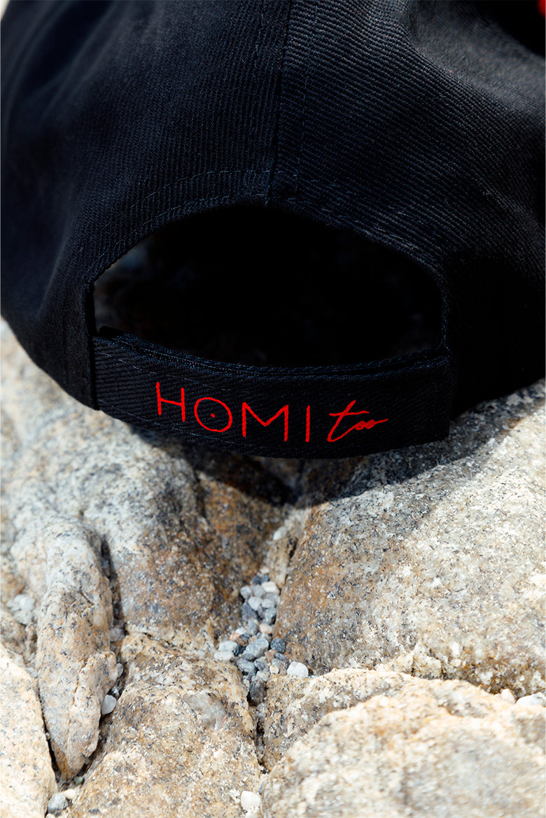 Close-up of the back of the black peaked cap showing the red "Homi Too" embroidery on the closure strap.