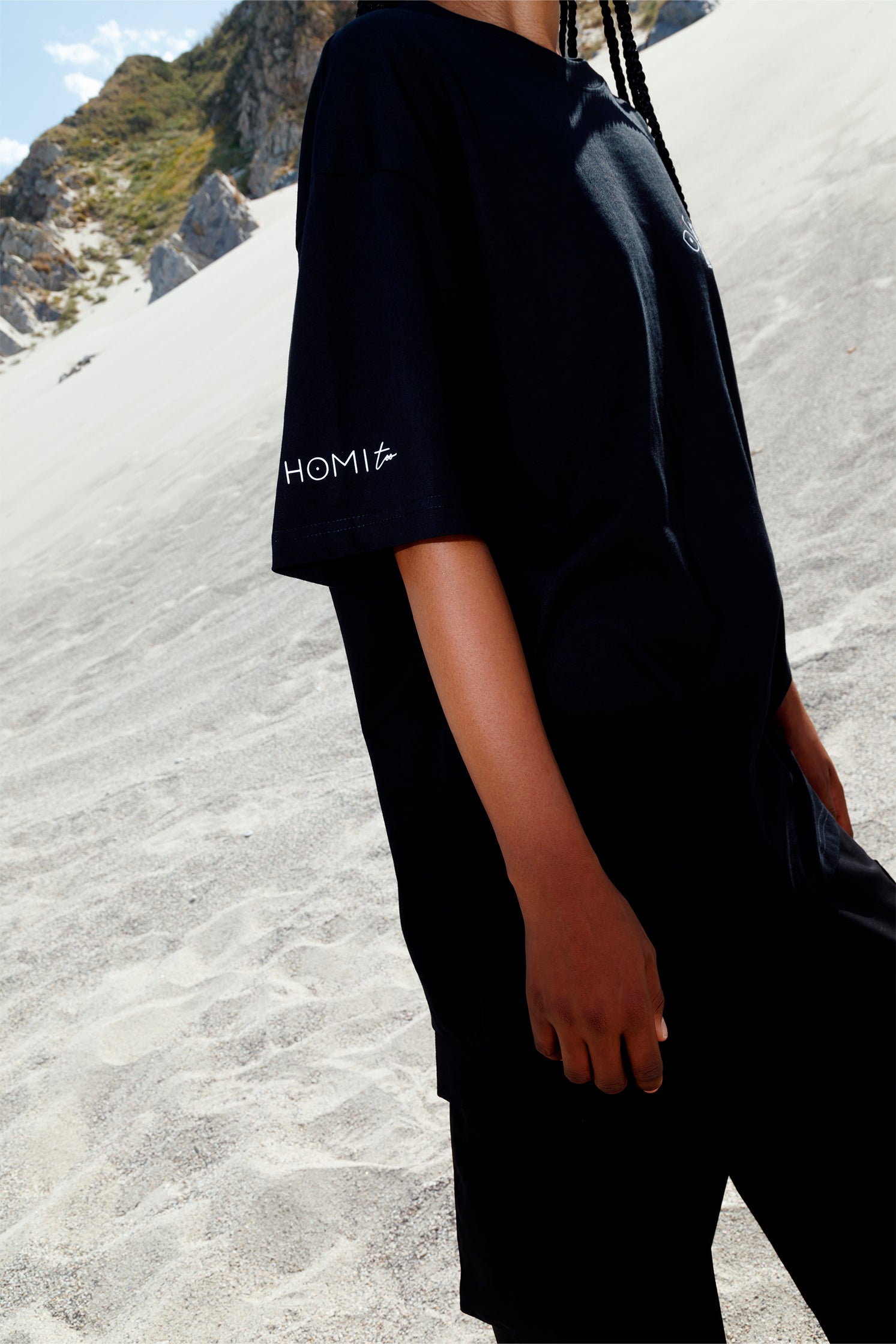 Close-up of the "Homi Too" print on the right sleeve of the black short-sleeved oversized t-shirt, highlighting clean lettering and refined placement.