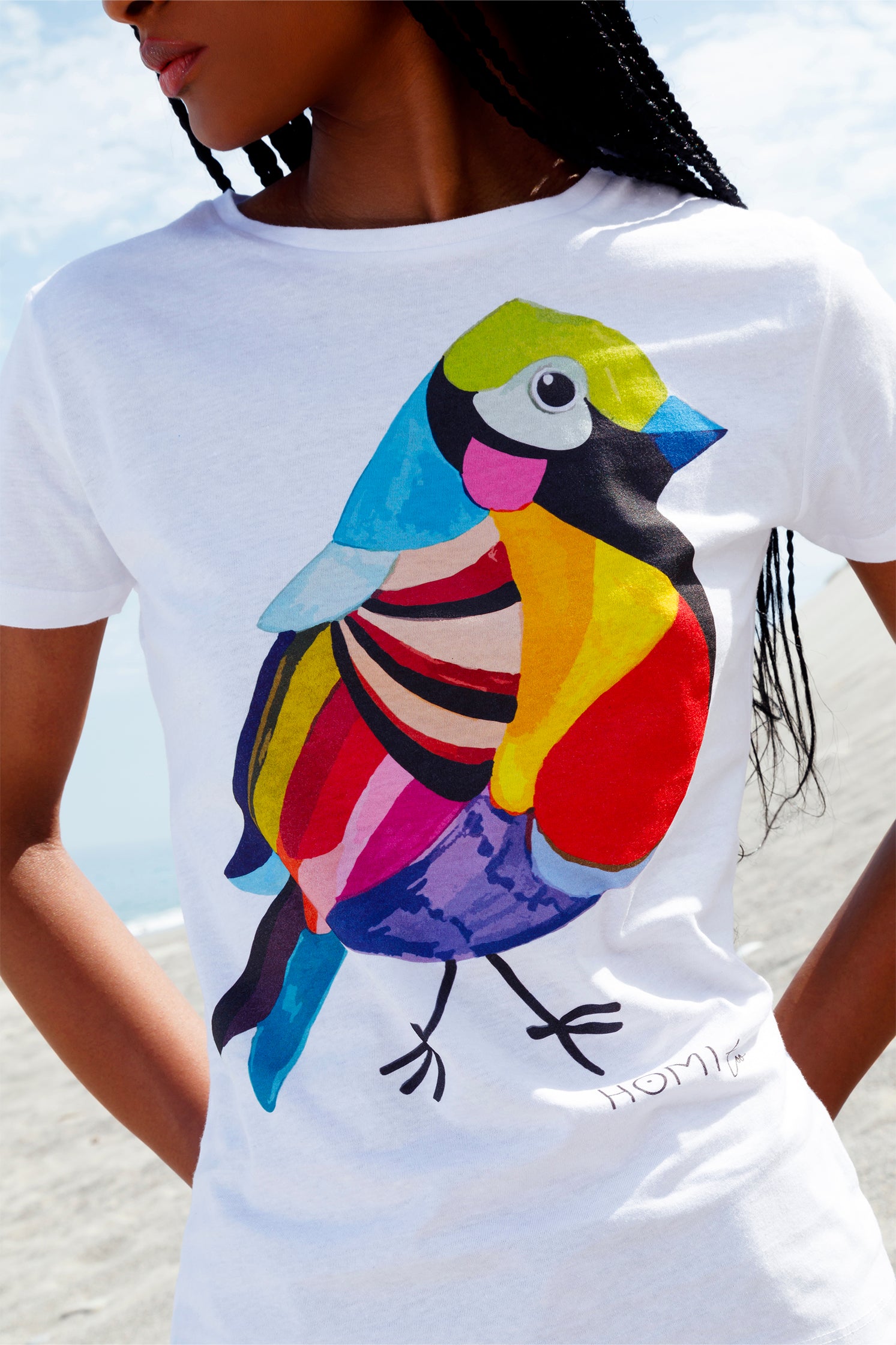 White slim women's t-shirt worn by a model, featuring a hand-painted colorful bird graphic on the front, "Homi Too" printed in small letters below the graphic on the left side, classic crew-neck, and short sleeves.