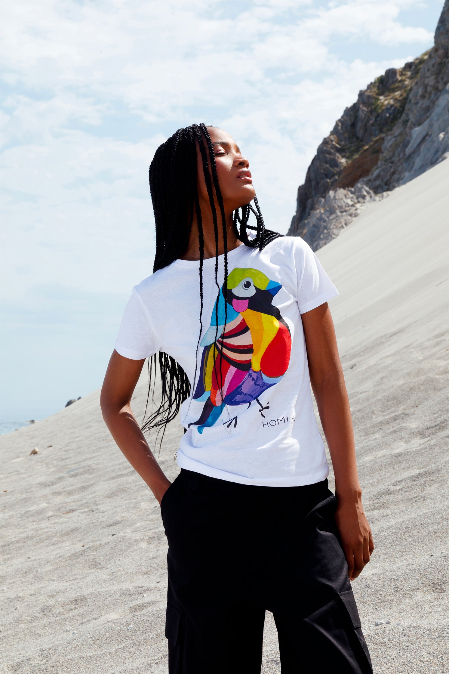 White slim women's t-shirt worn by a model, featuring a hand-painted colorful bird graphic on the front, "Homi Too" printed in small letters below the graphic on the left side, classic crew-neck, and short sleeves.