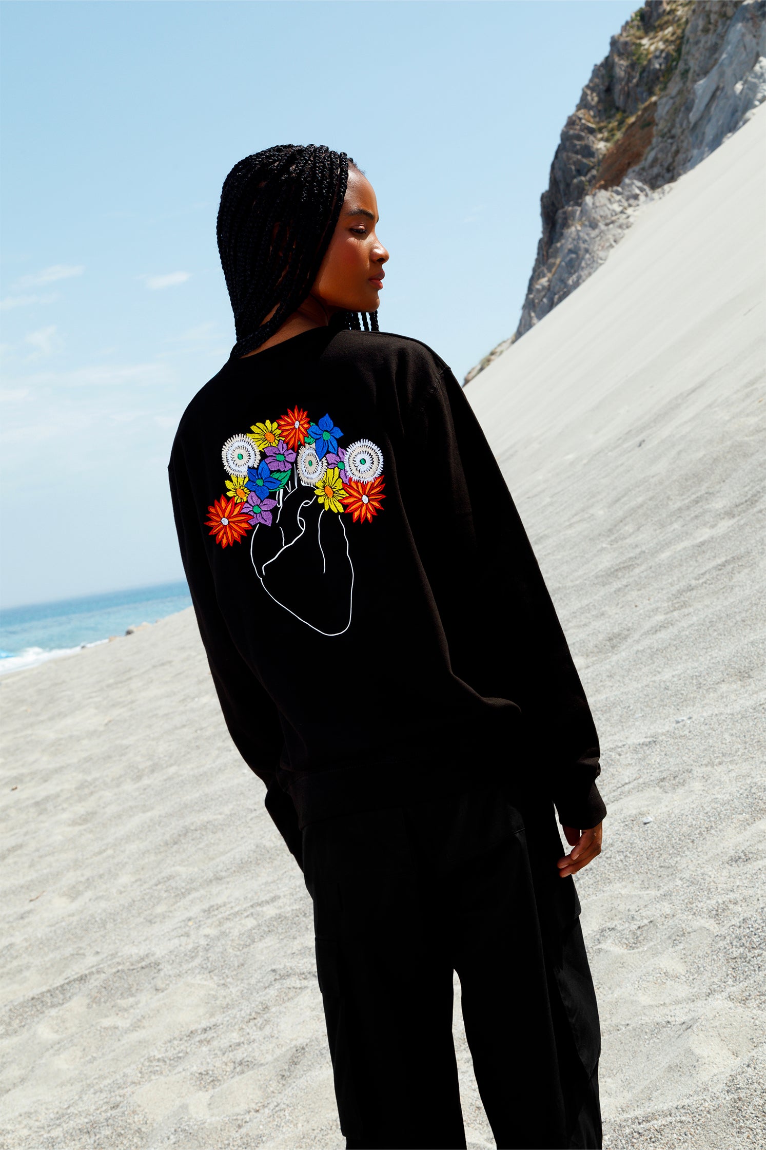 Back view of black oversized unisex sweatshirt worn by a model, showing the white heart embroidery with colorful flowers, crafted with artisanal precision.