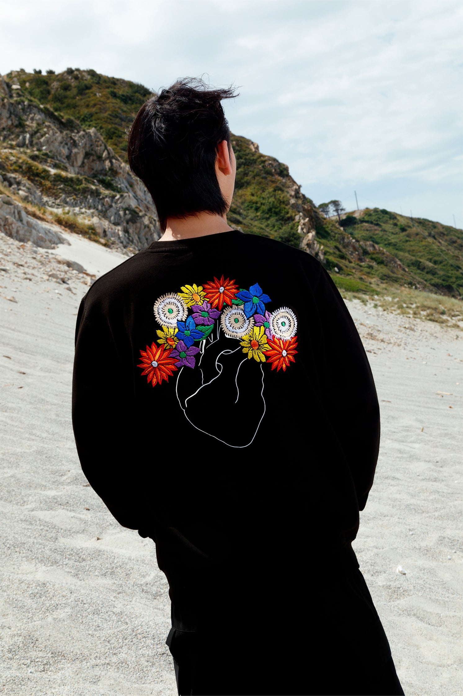 Back view of black oversized unisex sweatshirt worn by a model, showing the white heart embroidery with colorful flowers, crafted with artisanal precision.