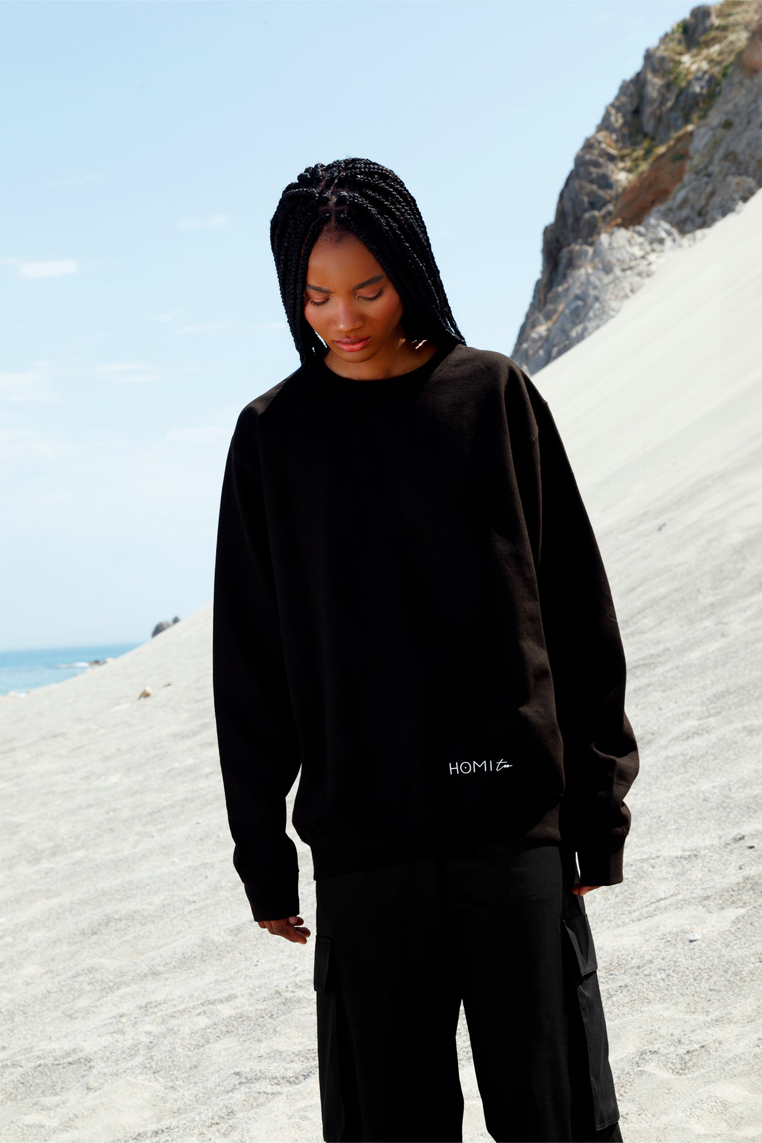 Black oversized unisex sweatshirt worn by a model, featuring a comfortable fit, with the white "Homi Too" logo printed on the front bottom left.