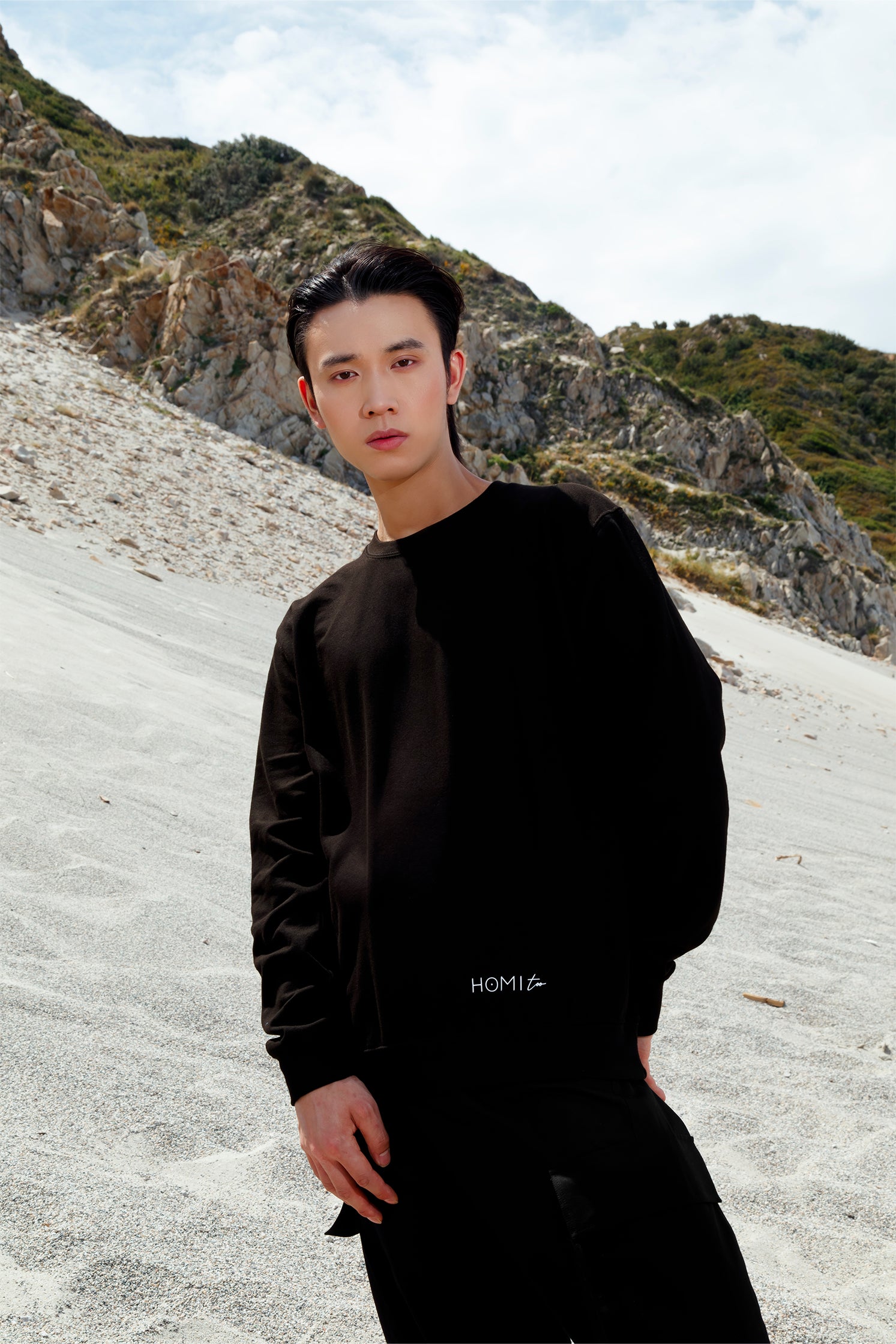 Black oversized unisex sweatshirt worn by a model, featuring a comfortable fit, with the white "Homi Too" logo printed on the front bottom left.