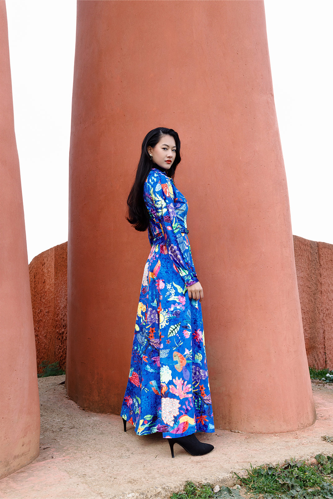 Side view of the long blue velvet dress worn by a model, featuring an exclusive artistic graphic, shirt collar, long sleeves, and fluid silhouette.