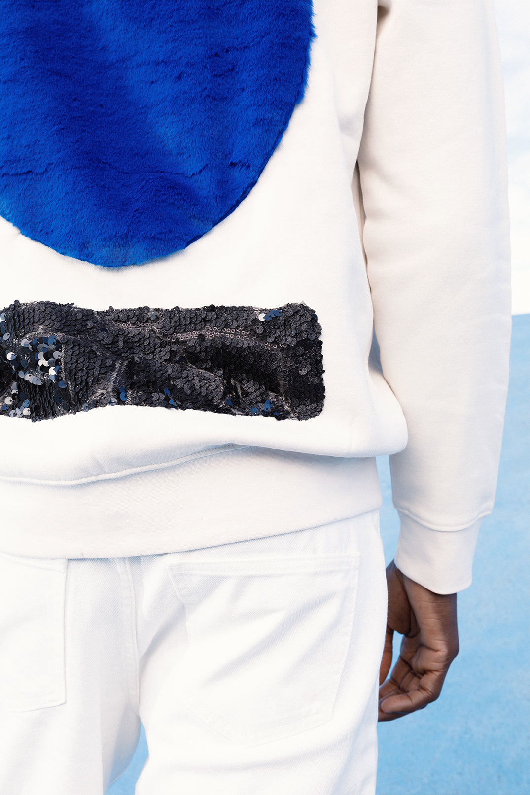 Close-up of the back of the white sweatshirt worn by a model, showing both the large soft blue faux fur circle and the lower strip of black sequins reflecting light.