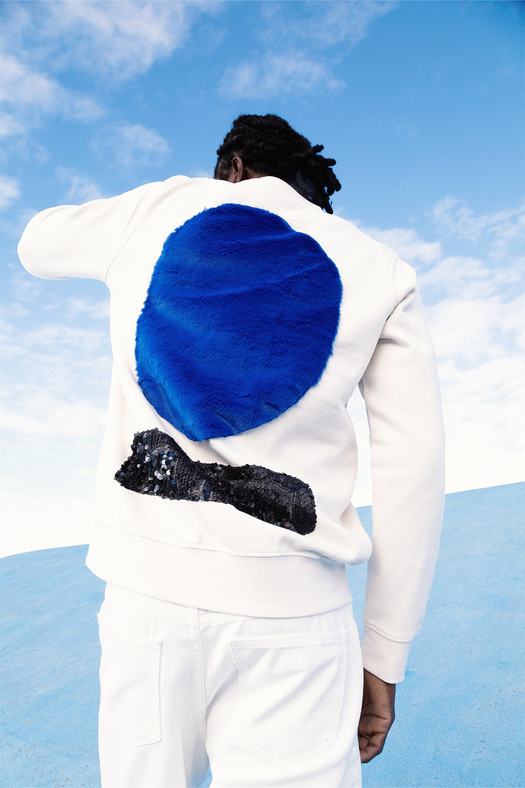 Back of the white sweatshirt worn by a model, with a large soft blue faux fur circle and a lower strip of black sequins reflecting light.