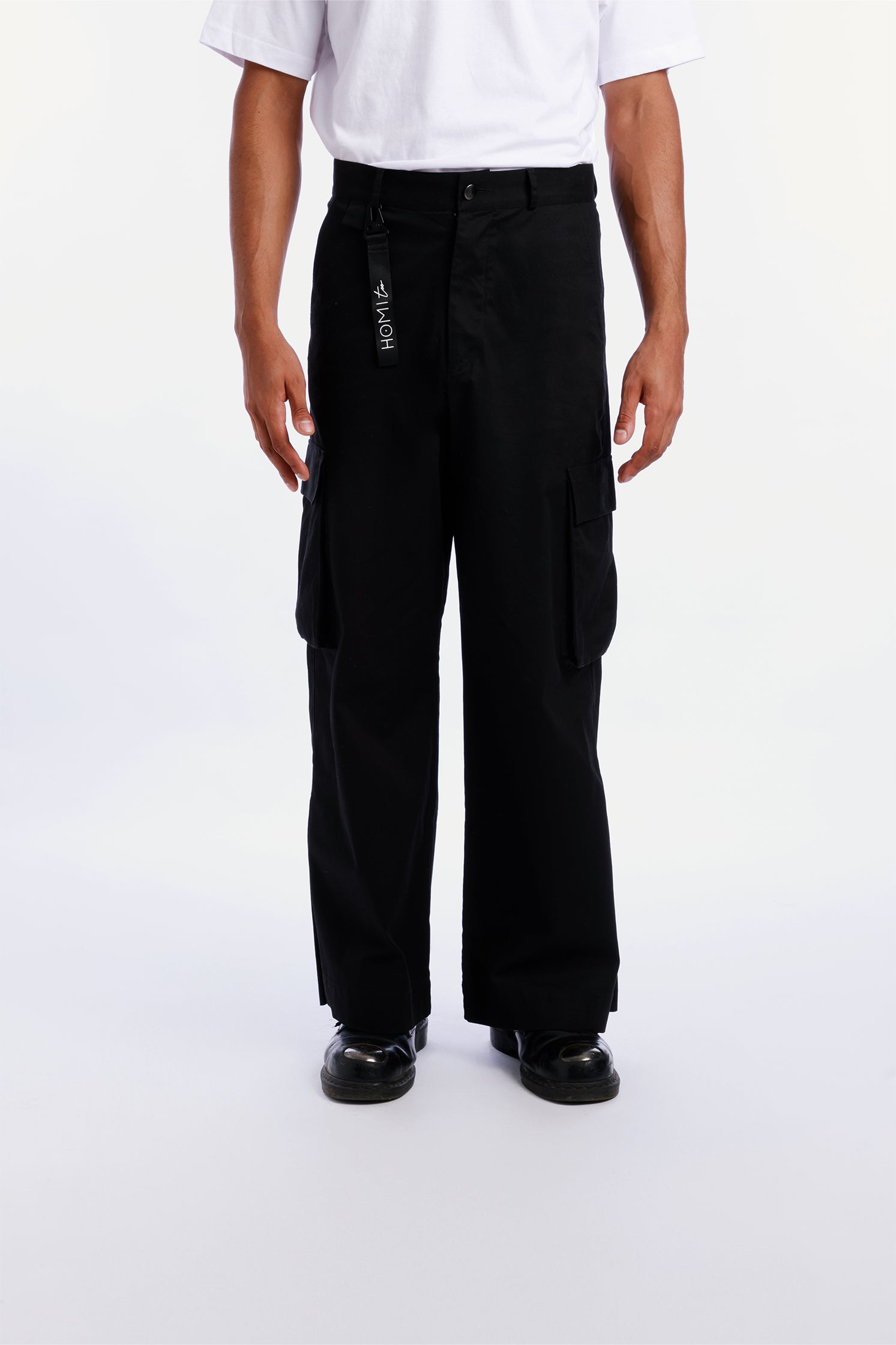 Unisex black cargo pants worn by a model with relaxed fit and side patch pockets, paired with a black keychain featuring the "Homi Too" logo in white lettering.
