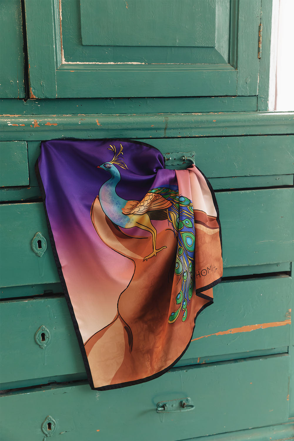 Square satin scarf with black border, featuring a colorful peacock perched on draped fabric against a vibrant purple, blue and golden background.