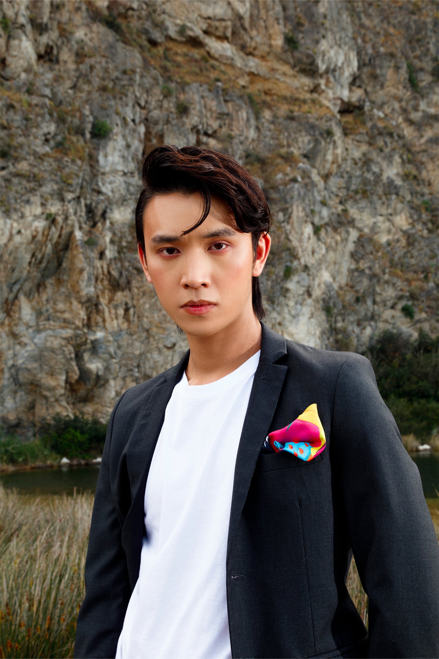 Silk scarf folded as a pocket square in a jacket worn by a model, showing the abstract multicolor geometric design on a blue-toned base.