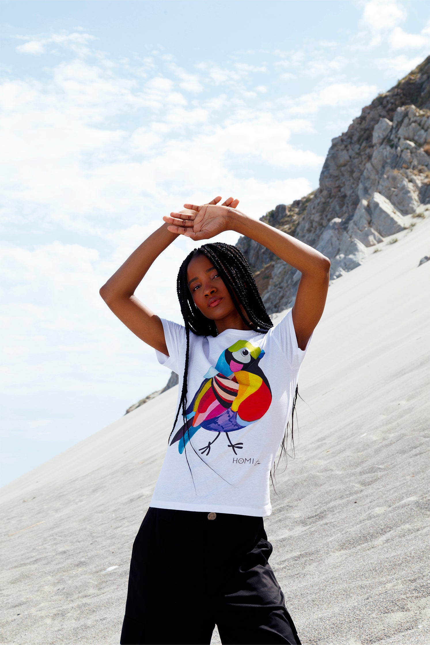 White slim women's t-shirt worn by a model, featuring a hand-painted colorful bird graphic on the front, "Homi Too" printed in small letters below the graphic on the left side, classic crew-neck, and short sleeves.