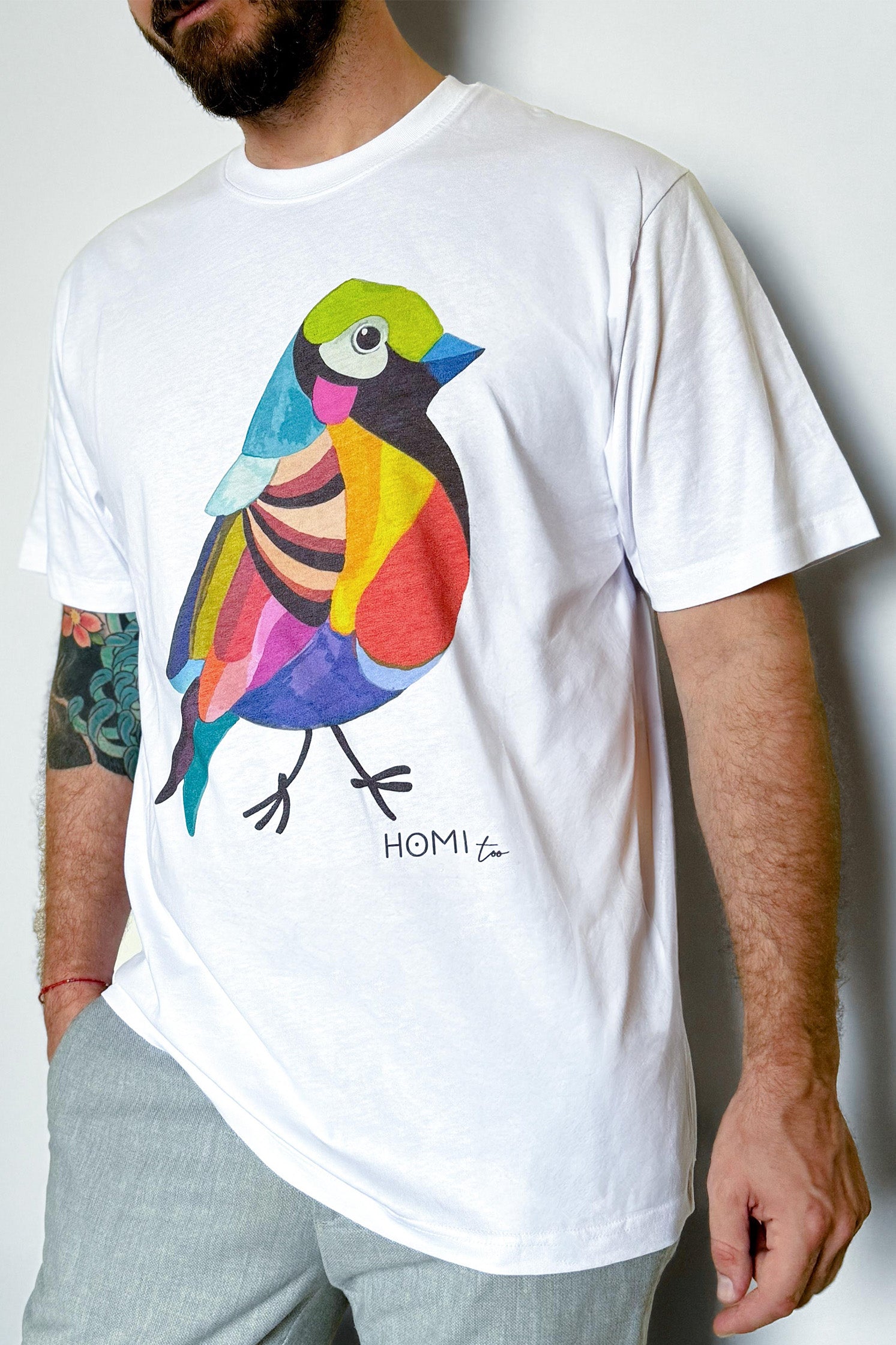 White oversized unisex t-shirt worn by a model, featuring a hand-painted colorful bird graphic on the front and "Homi Too" printed in small letters below the graphic on the left side.