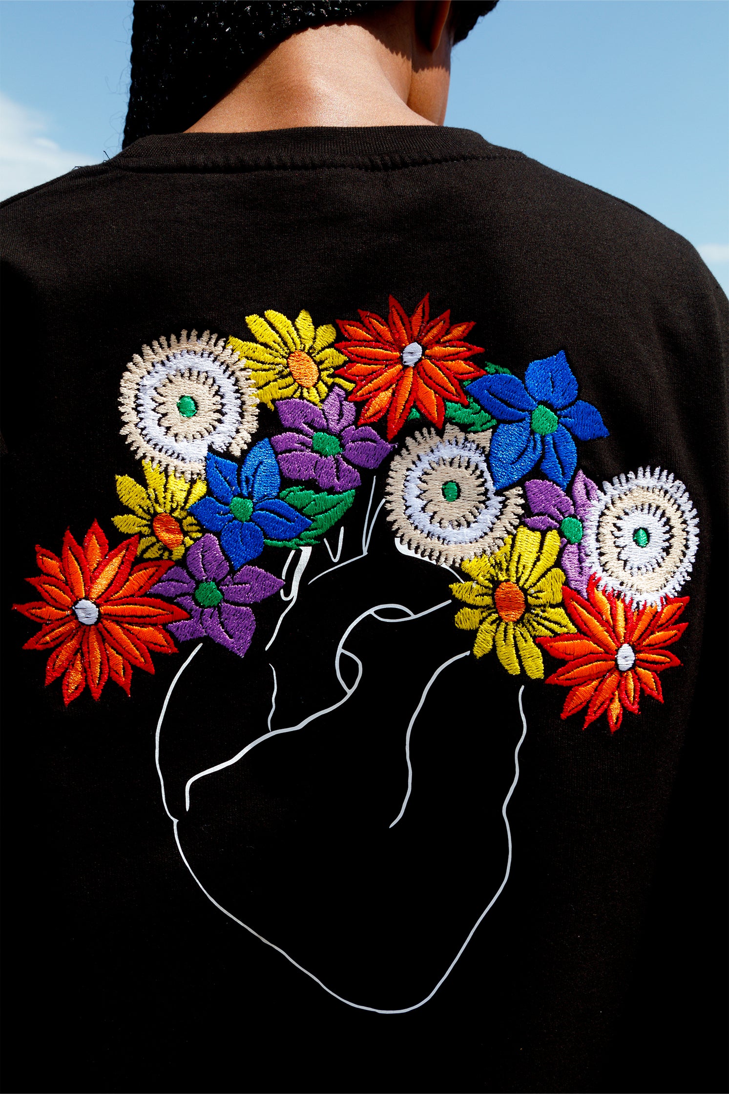Close-up of the white heart with colorful flowers embroidery on the back of the black oversized sweatshirt, highlighting intricate stitching and artisanal craftsmanship.