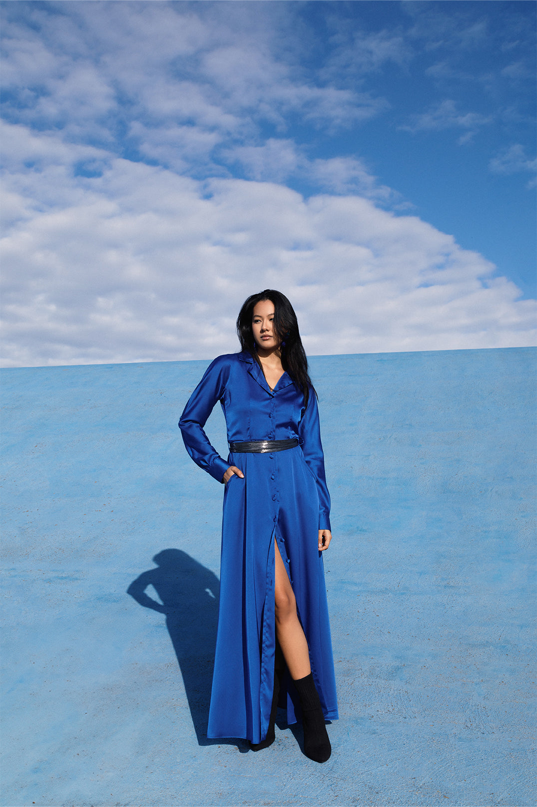 Elegant long royal blue dress worn by a model, with fluid silhouette, shirt collar, long sleeves.
