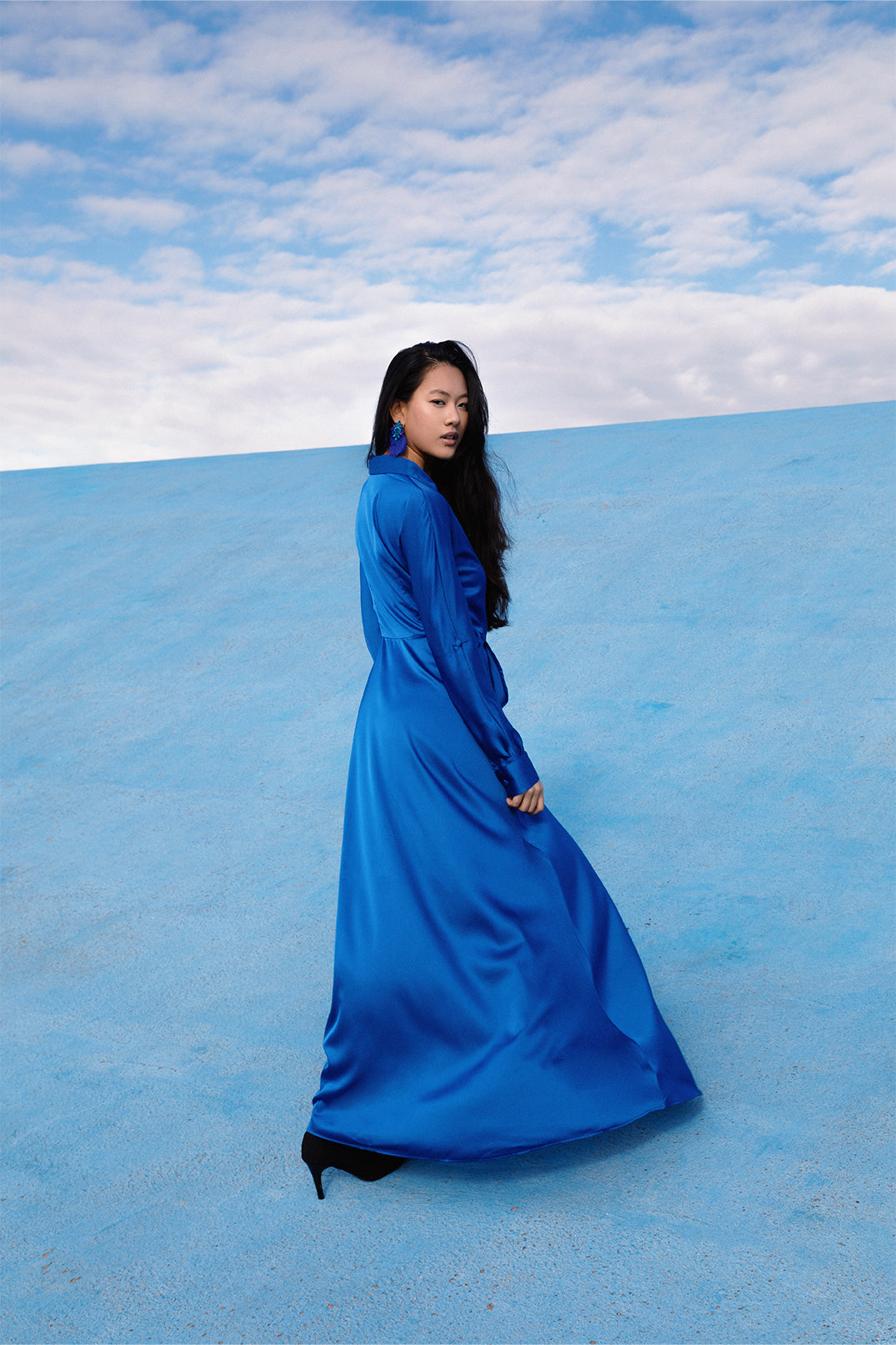 Back view of the elegant long royal blue dress worn by a model, with fluid silhouette, shirt collar, long sleeves.