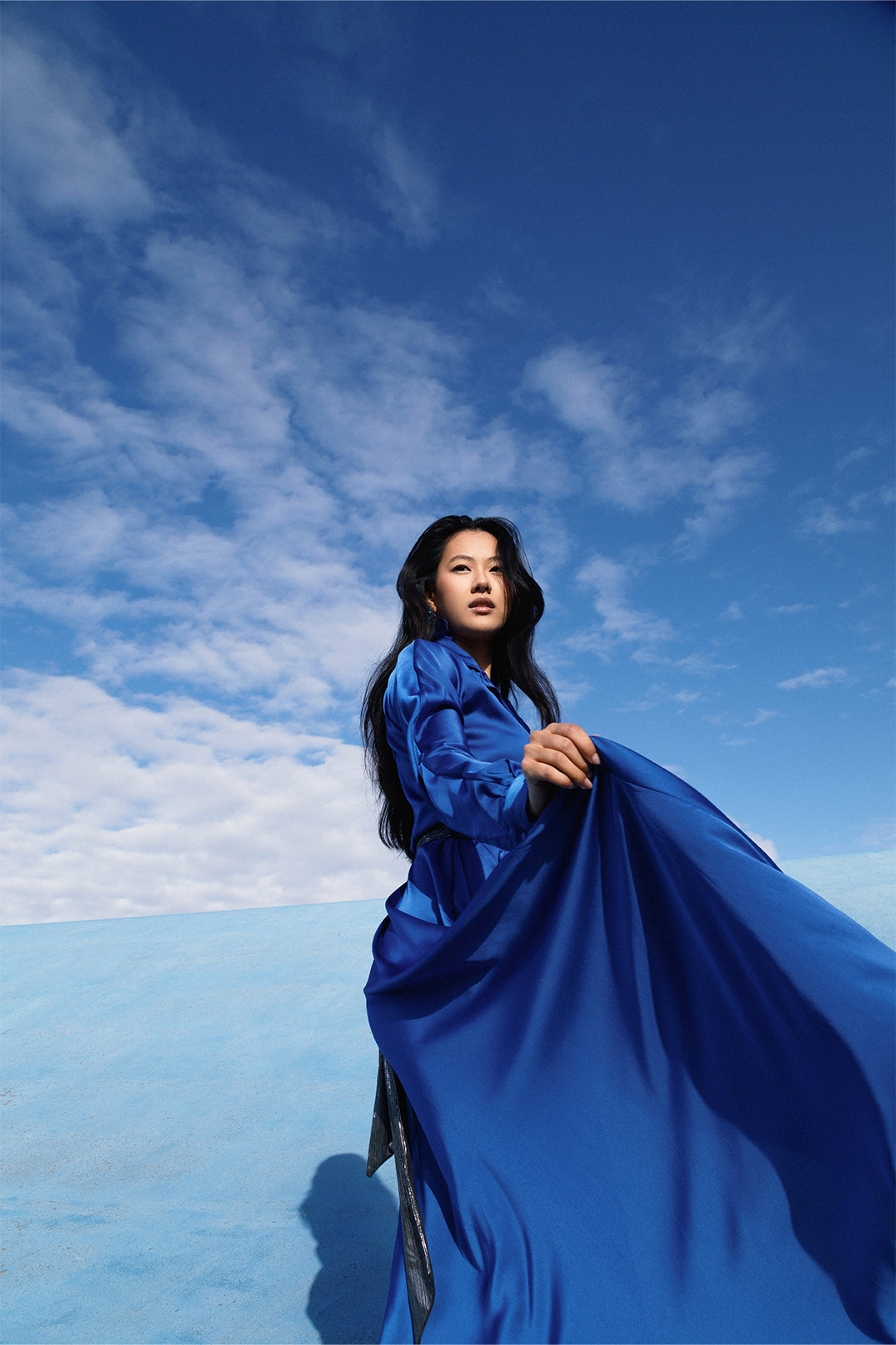 Elegant long royal blue dress worn by a model, with fluid silhouette, shirt collar, long sleeves.