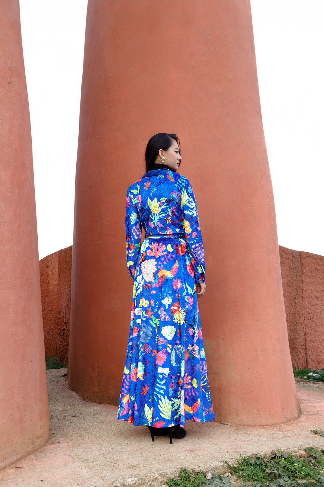 Back view of the long blue velvet dress worn by a model, featuring an exclusive artistic graphic, shirt collar, long sleeves, and fluid silhouette.