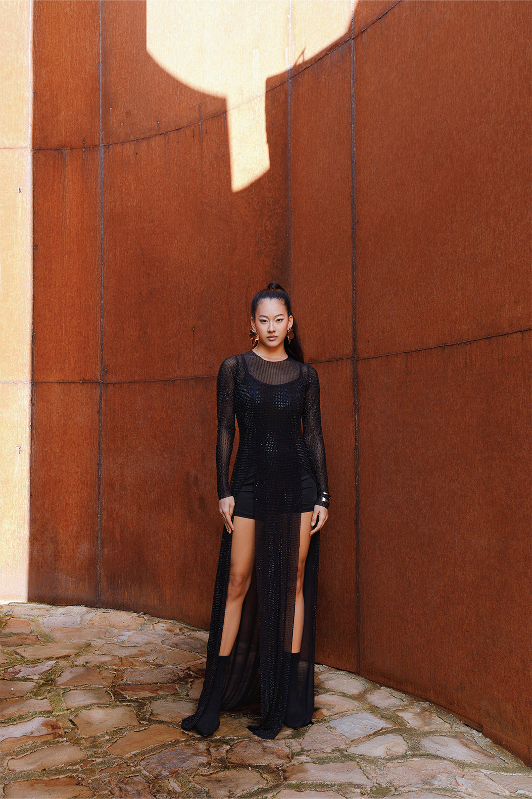 Long black dress worn by a model, featuring sheer details and sparkling rhinestones on lightweight fabric, flowing silhouette with side slits and sheer sleeves.