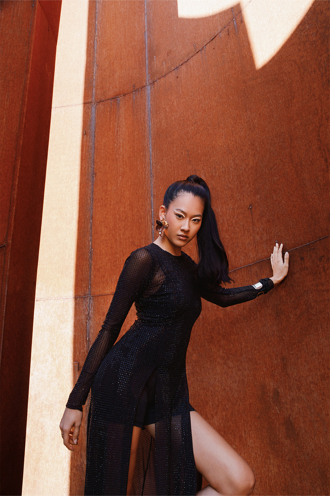 Long black dress worn by a model, featuring sheer details and sparkling rhinestones on lightweight fabric, flowing silhouette with side slits and sheer sleeves.