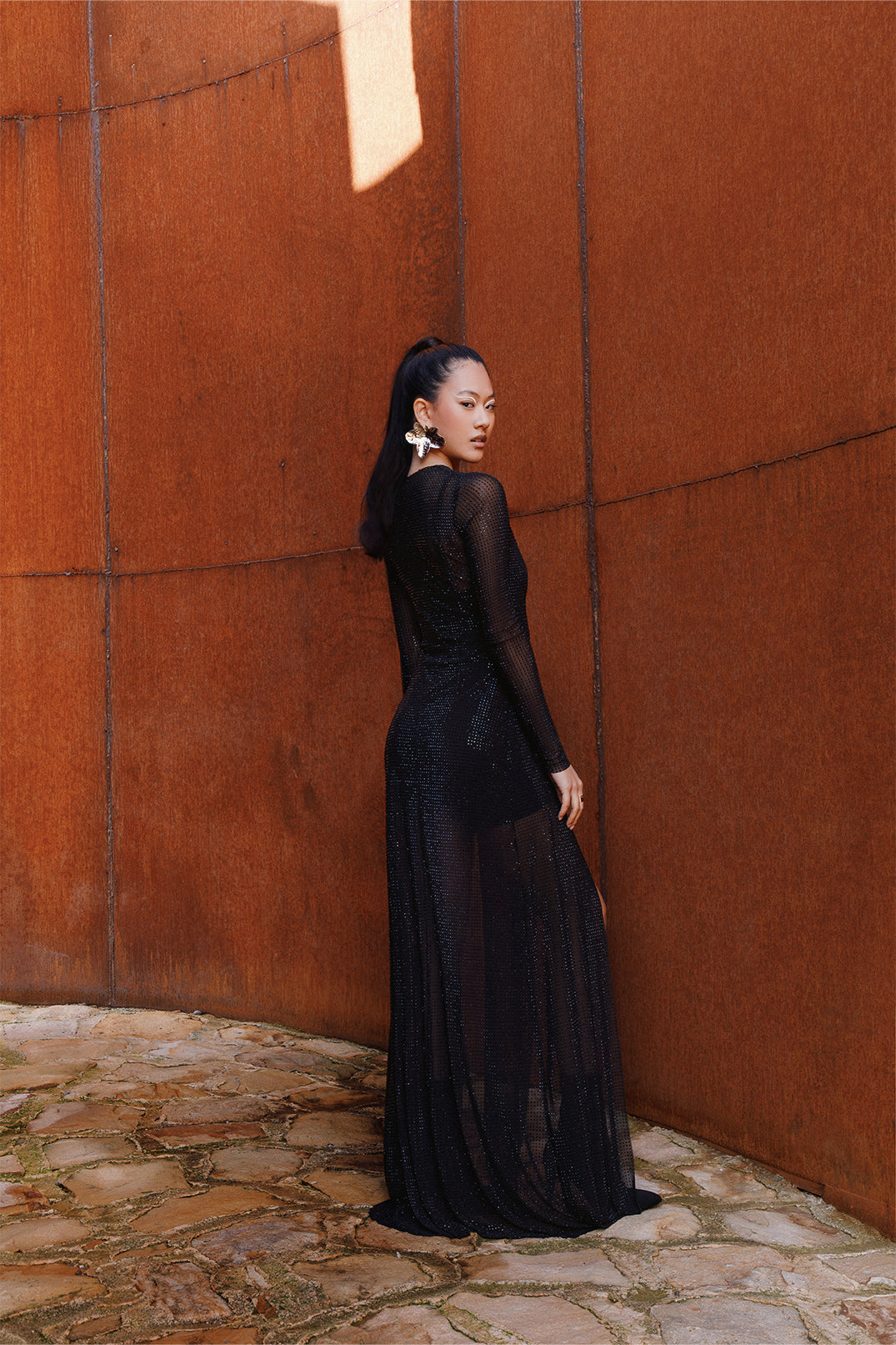 Back view of the long black dress worn by a model, featuring sheer details and sparkling rhinestones on lightweight fabric, flowing silhouette with side slits and sheer sleeves.