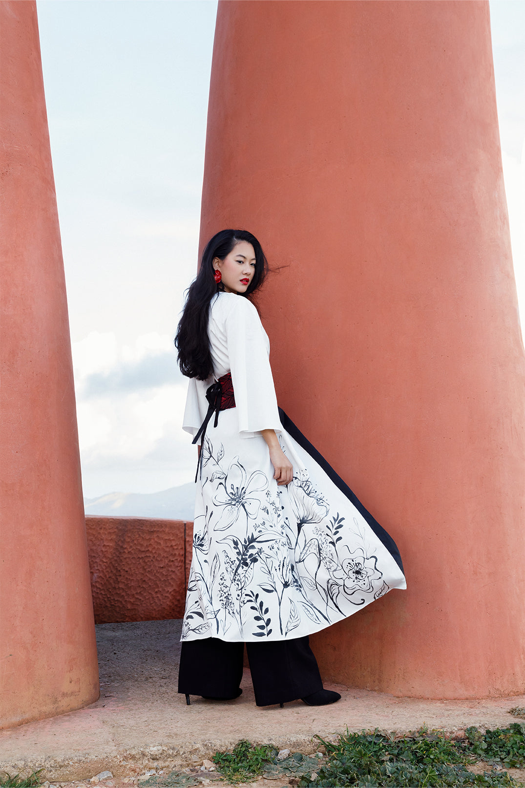 Back view of the long stretch white cotton kimono worn by a model, featuring elegant hand-painted floral illustration directly on the fabric, with a brocade belt at the waist for a fluid and sophisticated silhouette.
