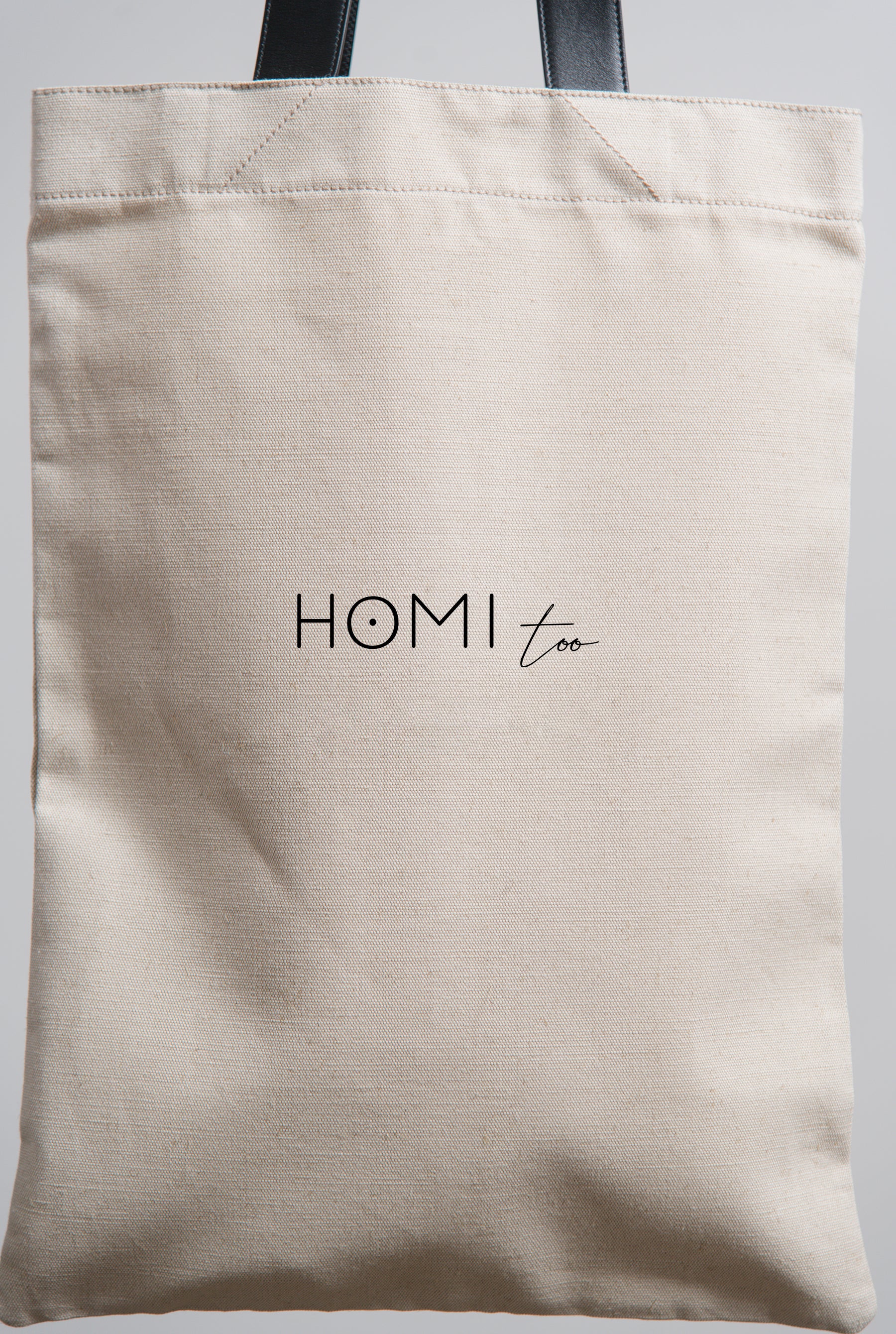 Back of the natural cotton shopper bag showing the 'Homi Too' logo centered, printed clearly on the fabric.