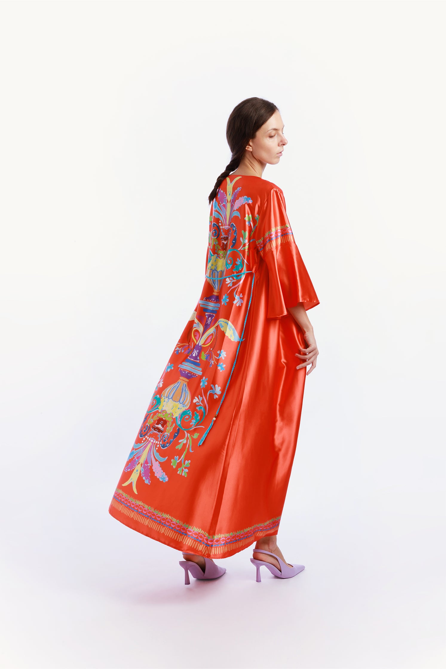Side view of the long caftan worn by a model, featuring an exclusive all-over print on the back inspired by majolica tiles and artistic decorations, with vibrant colors on a satin orange base, wide sleeves and soft fit for elegant comfort.