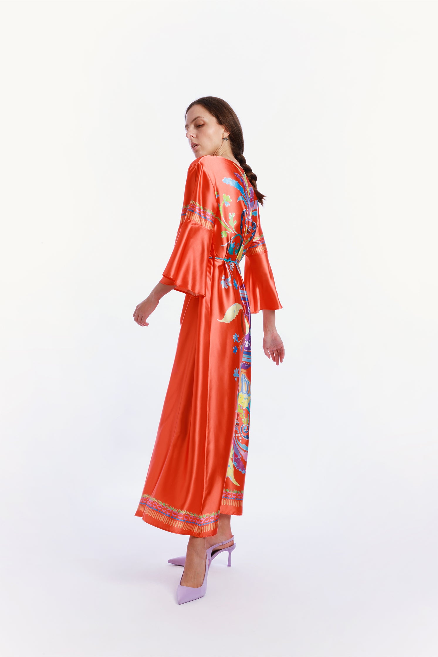 Side view of the long caftan worn by a model, featuring an exclusive all-over print on the back inspired by majolica tiles and artistic decorations, with vibrant colors on a satin orange base, wide sleeves and soft fit for elegant comfort.